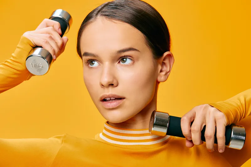 Female athlete performing upper body workout with dumbbells against a bright orange background, highlighting fitness, strength, focus, and athletic style in a modern studio.