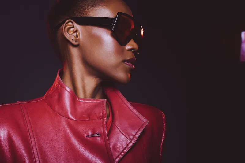 Portrait of confident African woman with dark skin wearing red leather jacket and large sunglasses. Studio lighting highlights her stylish look and strong facial profile in fashion concept.