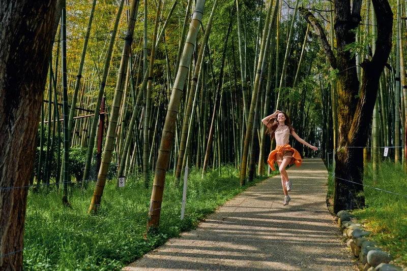 Bamboo forest, woman running on path in nature, joyful girl in orange dress skipping along sunlit walkway among tall bamboo trees, motion, freedom and outdoor leisure.