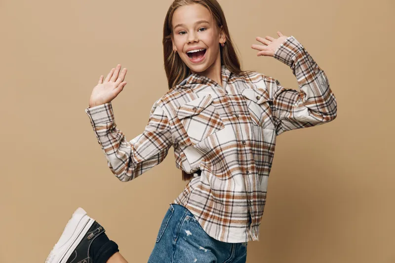 Smiling girl in plaid shirt and jeans jumping with raised hands on beige background, happy child expressing joy and excitement in casual outfit for outdoor or lifestyle.