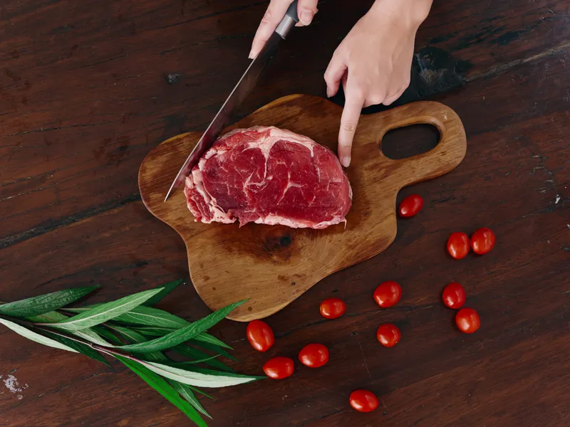 Женщина с ножом в руке нарезает свежее мясо для стейка, предназначенное для запекания на кухне