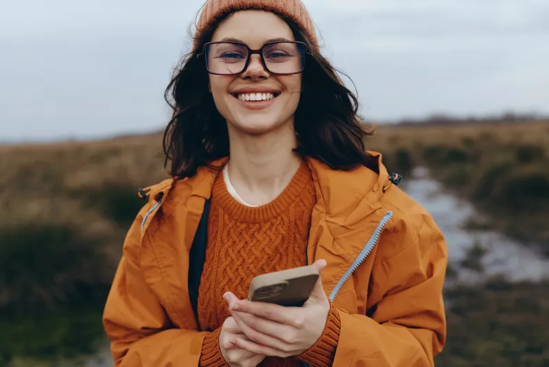 Woman lifestyle, smartphone outdoors autumn glasses beanie orange, smiling outdoors holding smartphone, casual sweater fashion, rural background, natural light, relaxed mood on trip