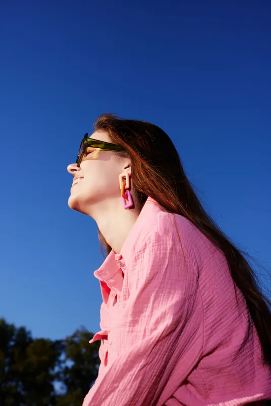 A smiling young woman wearing trendy pink clothing and colorful earrings against a clear blue sky, embodying summer fashion and positivity