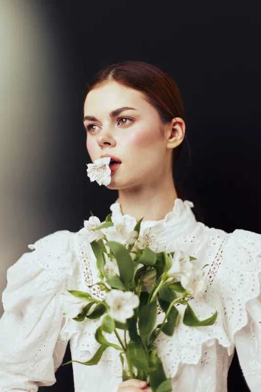 Elegant young woman in a white vintage dress holding flowers, with a flower in her mouth, showcasing serene emotions against a dark background