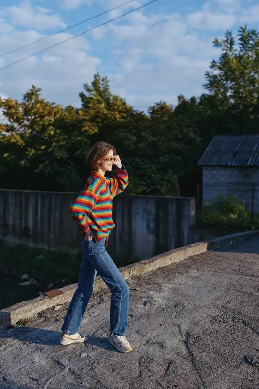 Woman in 20s wearing a colorful striped sweater and jeans walking on a rural rooftop, smiling with sunglasses in golden hour, relaxed casual outfit for outdoor lifestyle portrait.