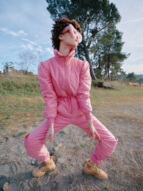 Outdoor scene with a woman in a pink jumpsuit striking a dynamic pose embracing bold movement amid a natural landscape trees and a bright blue sky