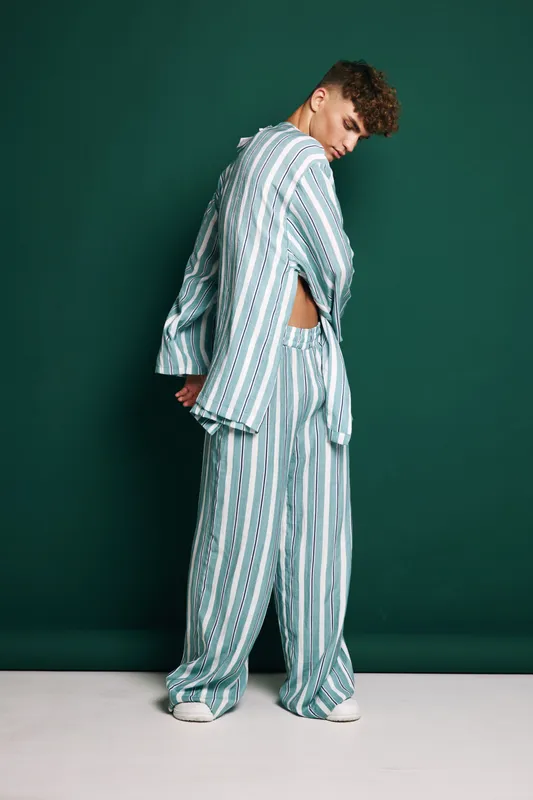 Young man in striped loose pajamas standing sideways against dark green background, casual pose with hands behind back and relaxed expression in studio setting.