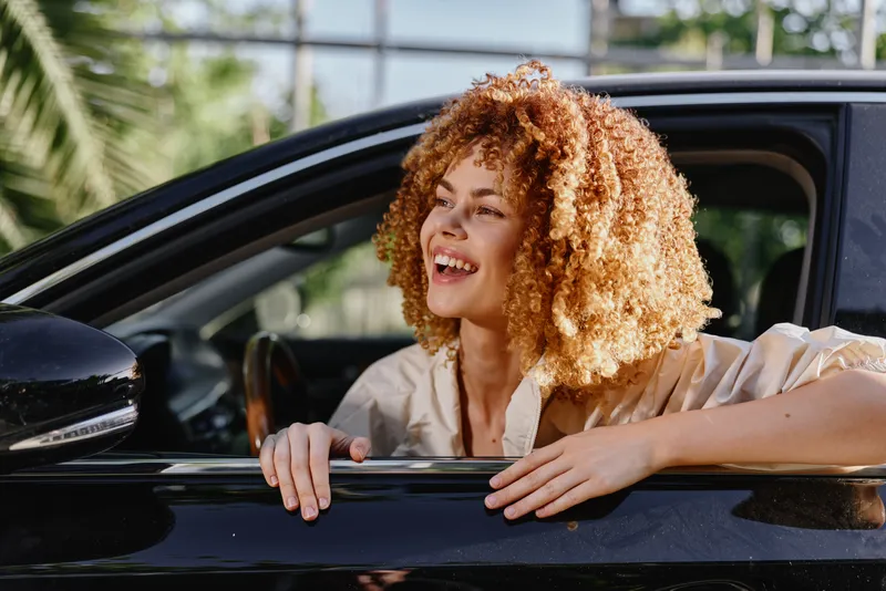 smiling woman with curly hair leaning out of car window in sunny outdoor setting, joyful, casual, natural light, morning mood