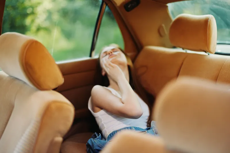 Young woman resting in a vintage car, wearing casual attire and expressing relaxation amidst a vibrant green background.