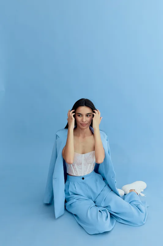 Young woman wearing blue suit and white lace top sitting on the floor against a blue background, styled with minimal makeup and casual elegance.
