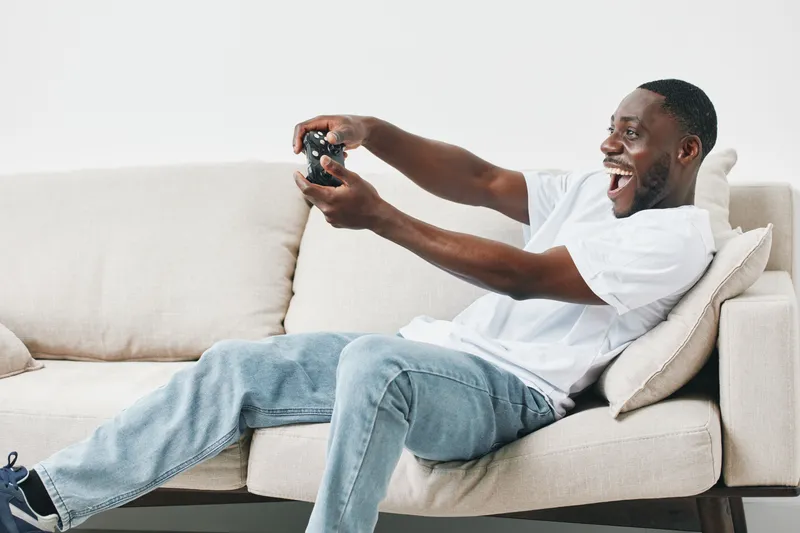 Happy young African man enjoying video games on the couch, wearing casual clothes, with a joyful expression in a modern living room setting