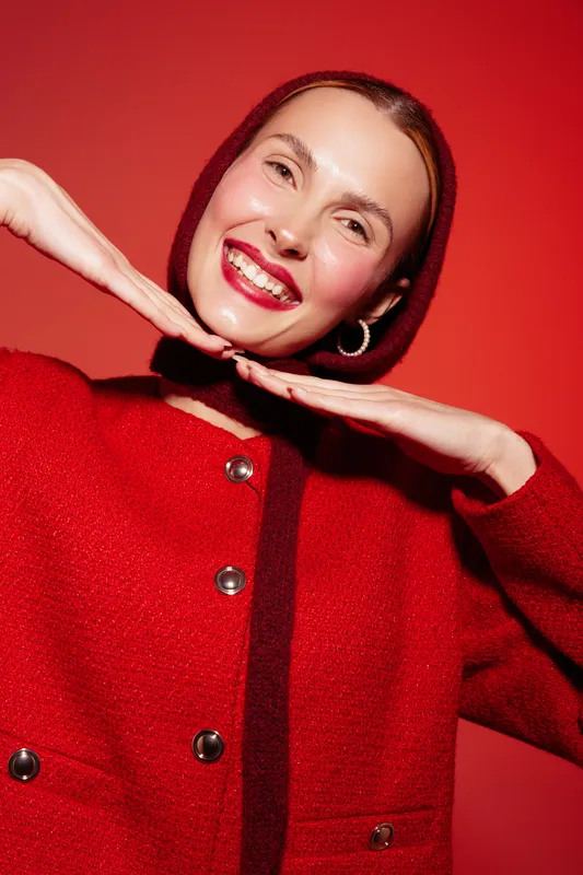 Smiling woman in red wool coat and headscarf poses with hands under chin on red background. Concept of fashion, winter style, happiness, and vibrant color contrast.