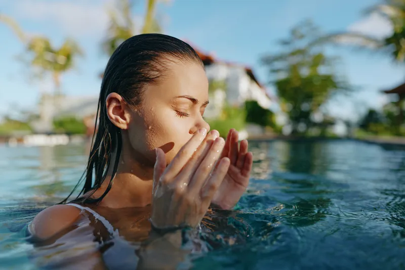 Young woman in a pool outdoors, hands cupping water near her face, sunlit moment, serene expression, relaxed posture, vibrant colors, fresh breeze, lifestyle mood