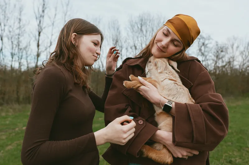 Lifestyle friendship outdoors women dog embrace park autumn casual affection, candid moment in cozy coats, serene outdoor scene with gentle smiles and warm tones.