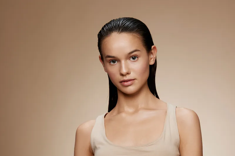 A serene young model with slicked back hair poses in a light beige top against a smooth, warm background, conveying confidence and calmness