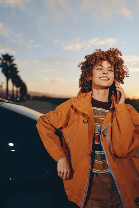 Woman outdoors, smiling at sunset, curly hair in warm light, wearing orange jacket and casual sweater, confident lifestyle travel mood, roadside background, soft evening breeze