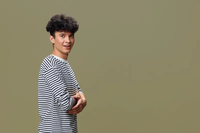 young man in striped shirt smiling confidently with arms crossed against plain olive background, casual style, curly hair, studio portrait