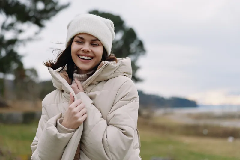Woman outdoor winter coat scarf and beanie smiles in a windy field, showing warmth and casual style. Natural light highlights a relaxed portrait with scenic nature setting and serene horizon.