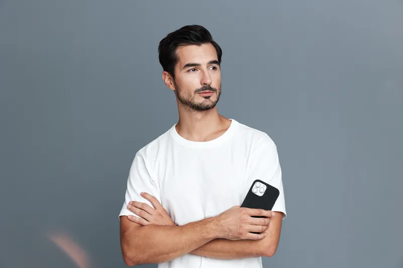 Handsome man in white t-shirt holds mobile phone while looking thoughtfully at camera, showcasing a casual, modern style in a neutral background. Lifestyle concept.