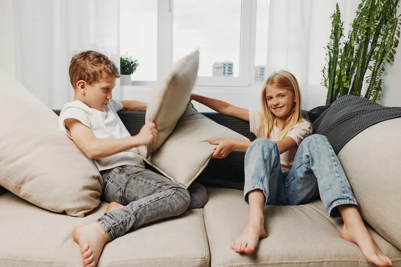 Two playful children having a pillow fight on a beige couch in a bright living room with large windows and green plants in the background. Concept of fun and childhood joy.