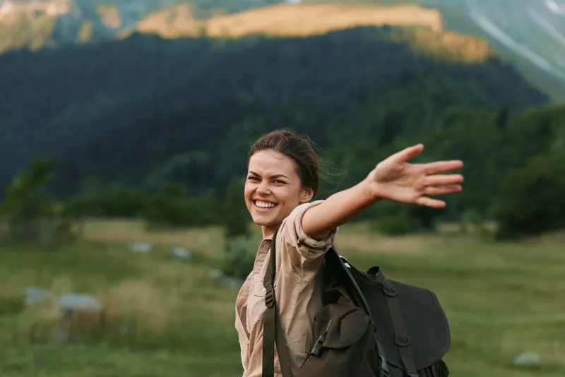 Woman lifestyle outdoors smiles while hiking with backpack in a grassy valley, enjoying nature and sunny moments, surrounded by mountains