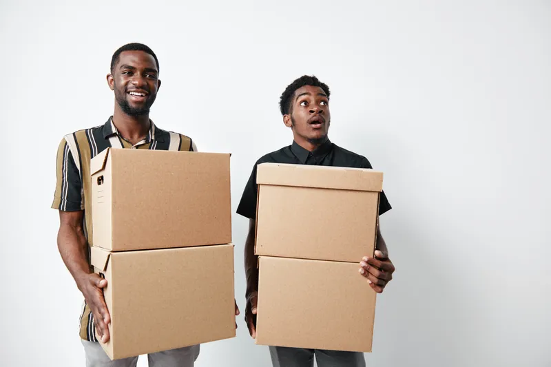 Two young black men holding cardboard boxes for moving or delivery, standing on white background. African ethnicity, dark skin, casual clothing and teamwork concept.