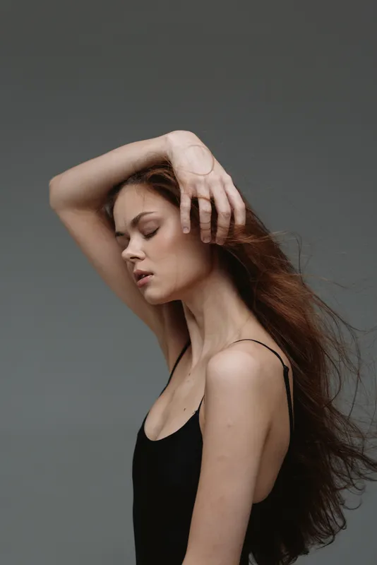 Elegant woman posing with eyes closed and hand on forehead, wearing black dress against gray background, peaceful mood.