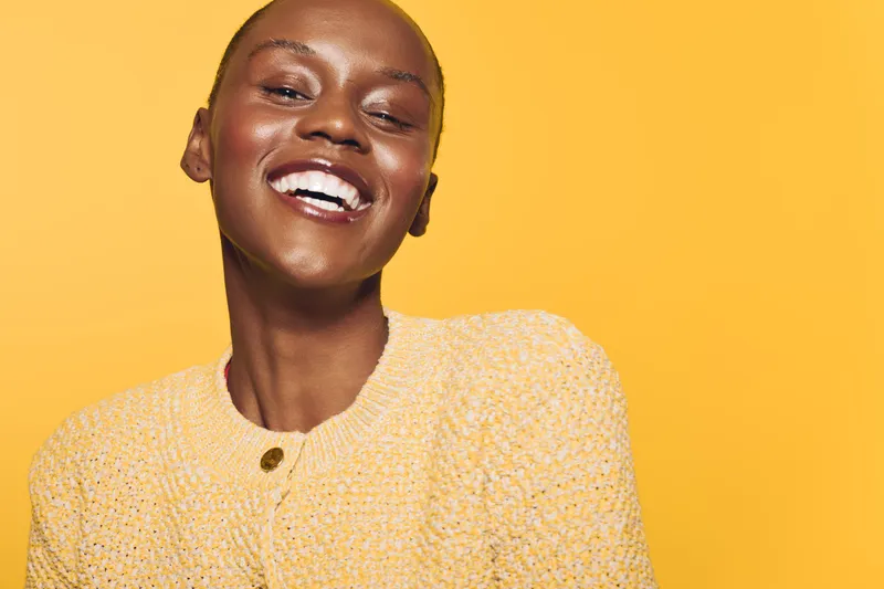 Happy African woman with dark skin smiling broadly, wearing a yellow knitted sweater against a yellow background. Concept of joy, beauty, fashion, and positive emotions in studio.