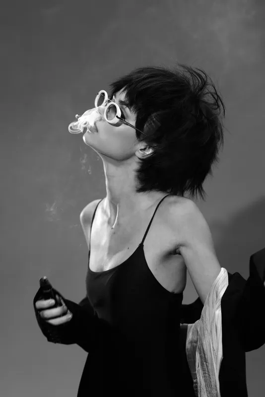Smiling woman in black dress and sunglasses blowing smoke in black and white artistic portrait