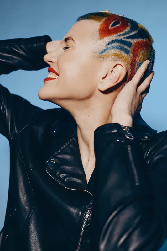Woman with colorful shaved hairstyle wearing a black leather jacket, smiling with eyes closed, showing confidence and edgy fashion style on blue background.