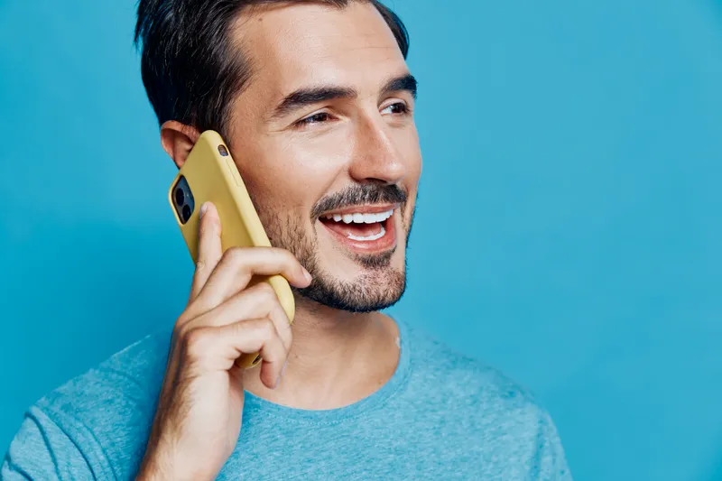 Smiling man with short dark hair wearing a blue shirt talking on a yellow smartphone against a vibrant blue background. People lifestyle concept.