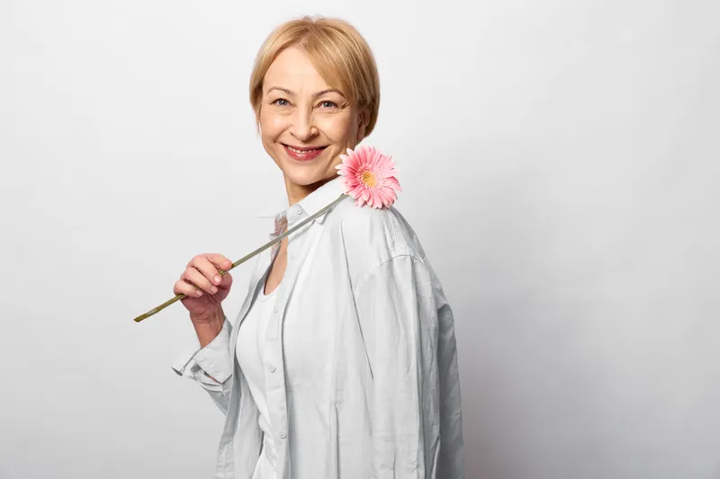Woman holding pink flower, studio portrait with gentle smile, light gray shirt, calm expression, soft background, spring mood, minimal composition, relaxed pose indoors