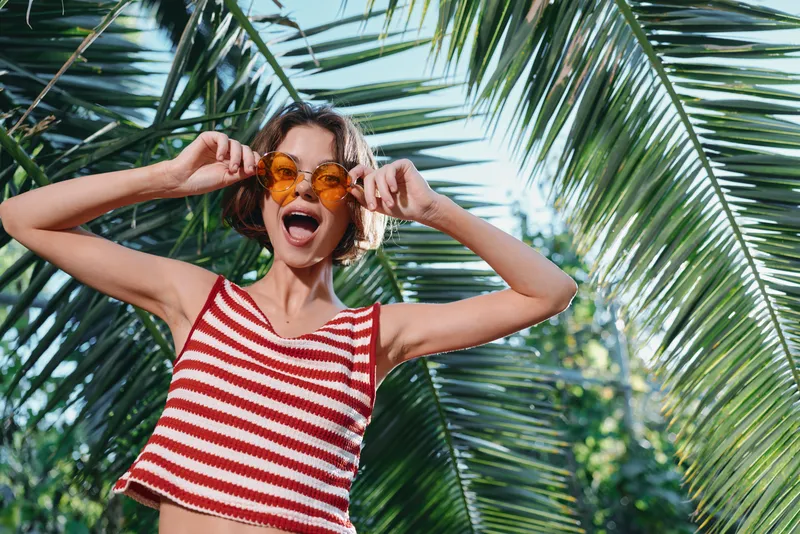 A cheerful young woman wearing a striped red and white top and round orange sunglasses poses playfully among lush palm leaves under bright sunlight.