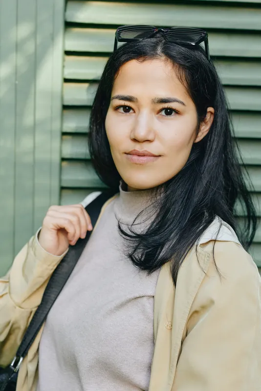 Casual outdoor photo of a woman wearing a beige jacket and sunglasses on her head, standing by a green backdrop, confident and relaxed, suitable for fashion and lifestyle campaigns