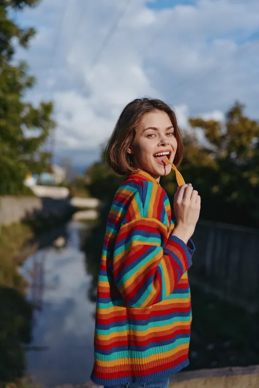 Woman in a colorful striped sweater smiling and eating a snack outdoors by a canal, young adult leaning on railing in casual outfit for playful lifestyle portrait.