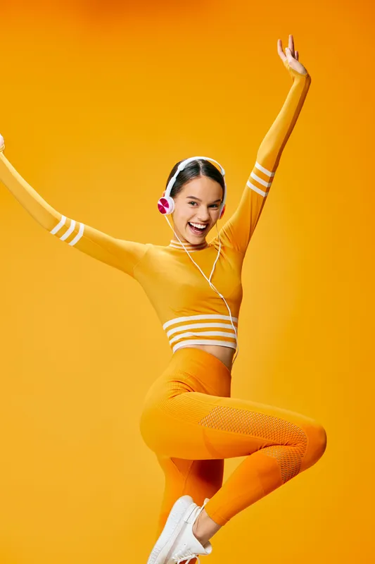 Energetic woman in vibrant orange athletic wear performs a high leap, showing balance and strength in a bright studio, a dynamic fitness shot with bold pose and joyful energy.