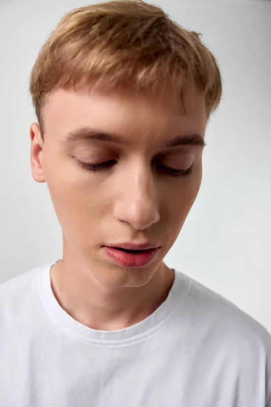 Closeup of a young man with downcast gaze, wearing a white shirt, conveying calm, contemplation, and gentle serenity in a minimal studio setting