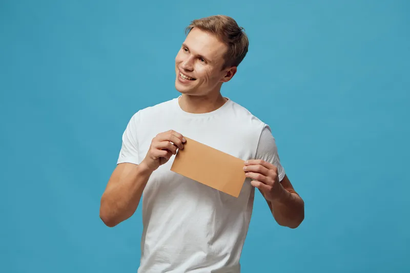 Smiling young caucasian man wearing white t-shirt hold kraft envelope look aside isolated on blue background mental health and gadgets concept