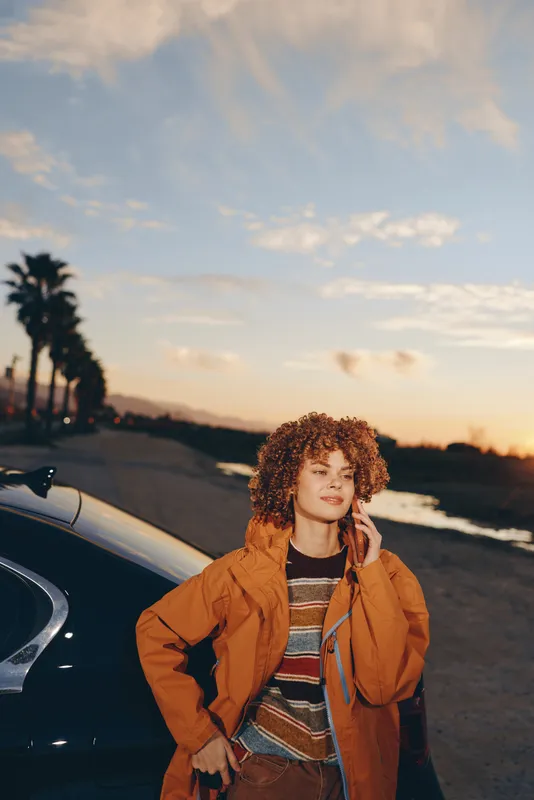 Woman lifestyle portrait outdoors near a car on a road at sunset, curly hair and smiling face, wearing a jacket, warm evening light, travel mood, calm roadside background