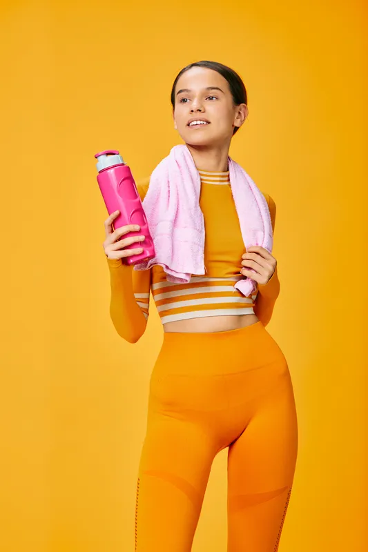 Active woman in bright orange fitness wear stands against a vivid orange backdrop, holding a pink water bottle with a towel draped around her neck, ready for workout.