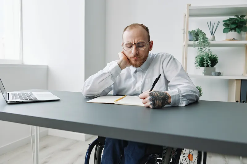 Мужчина в инвалидной коляске, бизнесмен, работает в офисе за ноутбуком, записывая план в блокнот