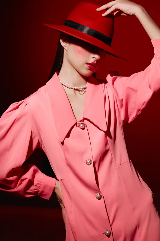Fashion model in pink blousy dress and red hat posing dramatically with hand raised over her face. Elegant style and makeup in creative beauty concept.