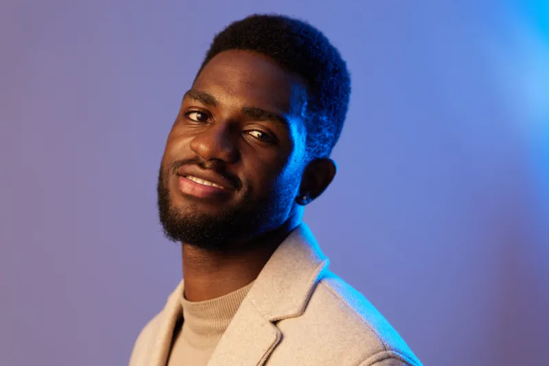 Confident young Black man in stylish beige coat, smiling against a soft blue background, showcasing modern fashion and charisma in a vibrant studio setting