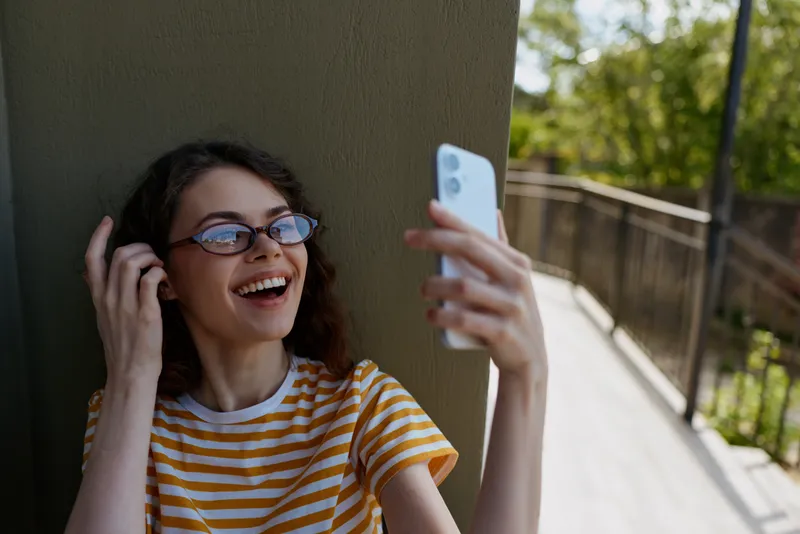 A young woman with a striped tee and glasses smiles while taking a selfie on a sunlit balcony, capturing a casual lifestyle moment with her phone.