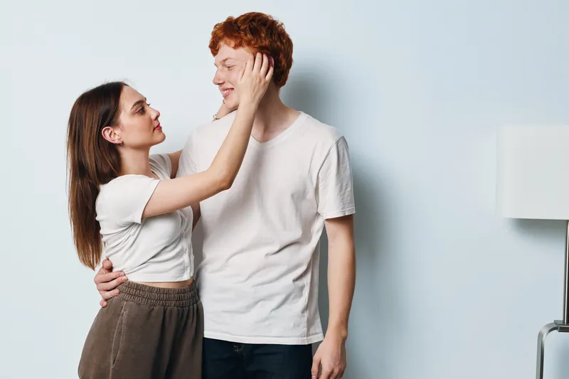 Couple embracing indoors, woman touching man's face with smile in casual clothes near lamp and light background, showing affection and intimacy in relationship.
