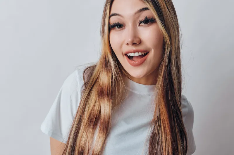 Young woman with long hair smiles happily wearing white t shirt studio portrait on plain background. Positive female model with natural makeup and straight hair indoors casual style.