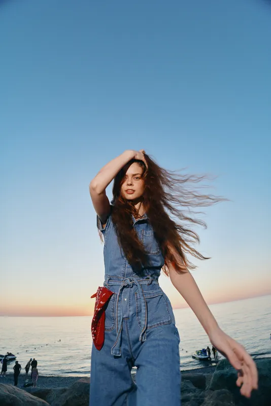 lifestyle woman in modern boho-western denim street style with flowing hair at seaside during sunset, film color tones enhance natural and relaxed vibe