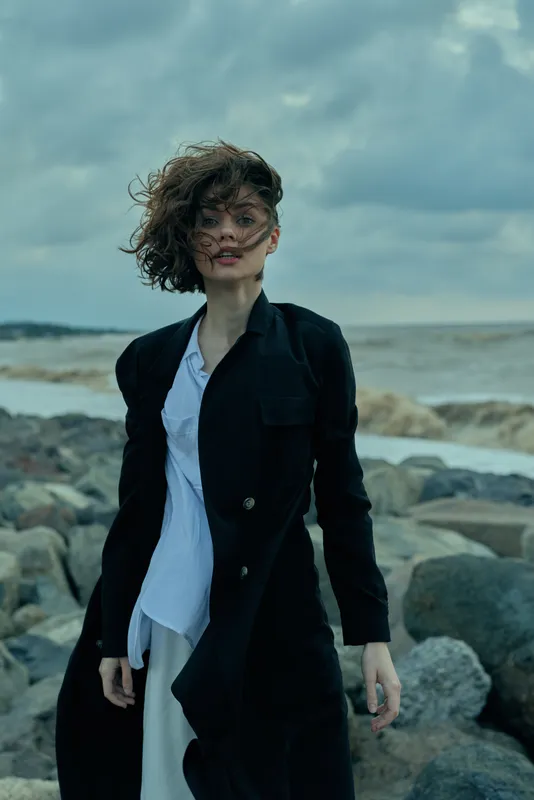 Windswept woman stands on a rocky coast in a long black coat and white shirt, dramatic sea backdrop and cloudy sky, a cinematic fashion moment with movement and strength.