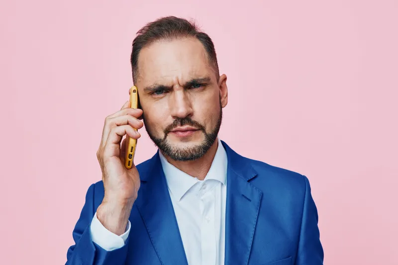 A professional man on a pink background speaks on a smartphone, projecting confidence and focus. The isolated colored backdrop emphasizes his business poise and serious demeanor.