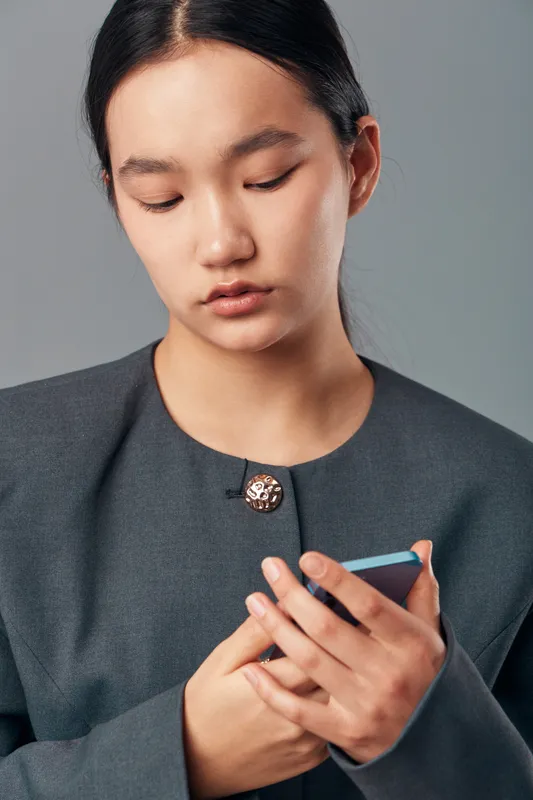 Young woman in gray blazer looking down at smartphone with focused expression, holding mobile device with both hands on plain gray background in studio setting.